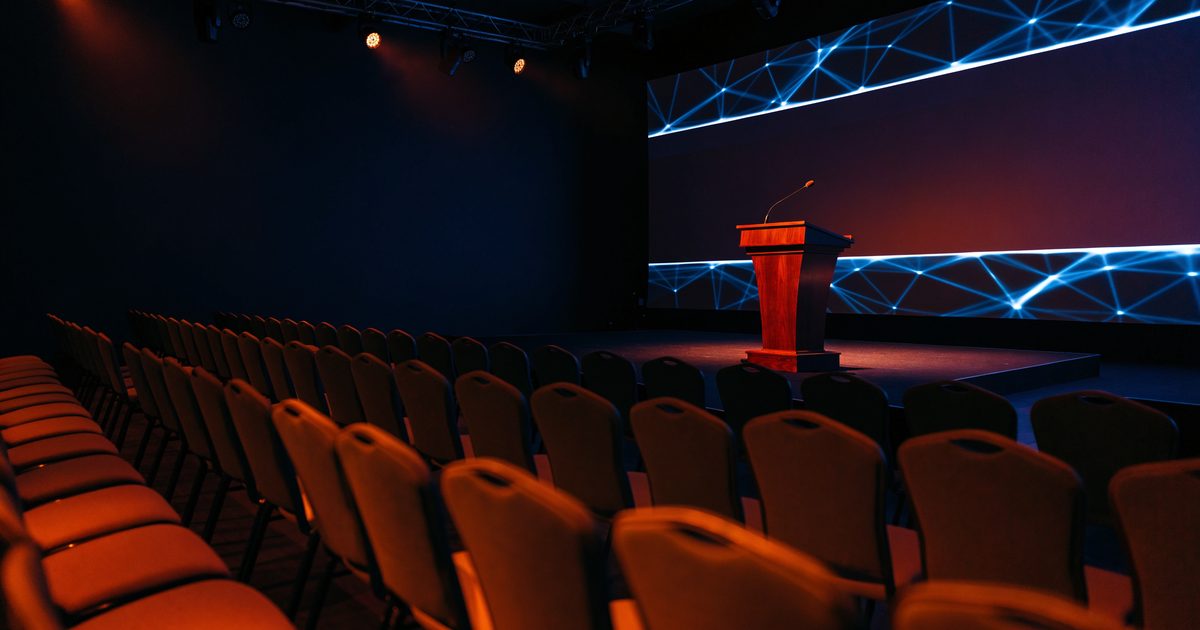 An empty conference ballroom before a keynote, with a lectern and microphone on stage, a glowing blue network pattern on the screen and rows of empty chairs.