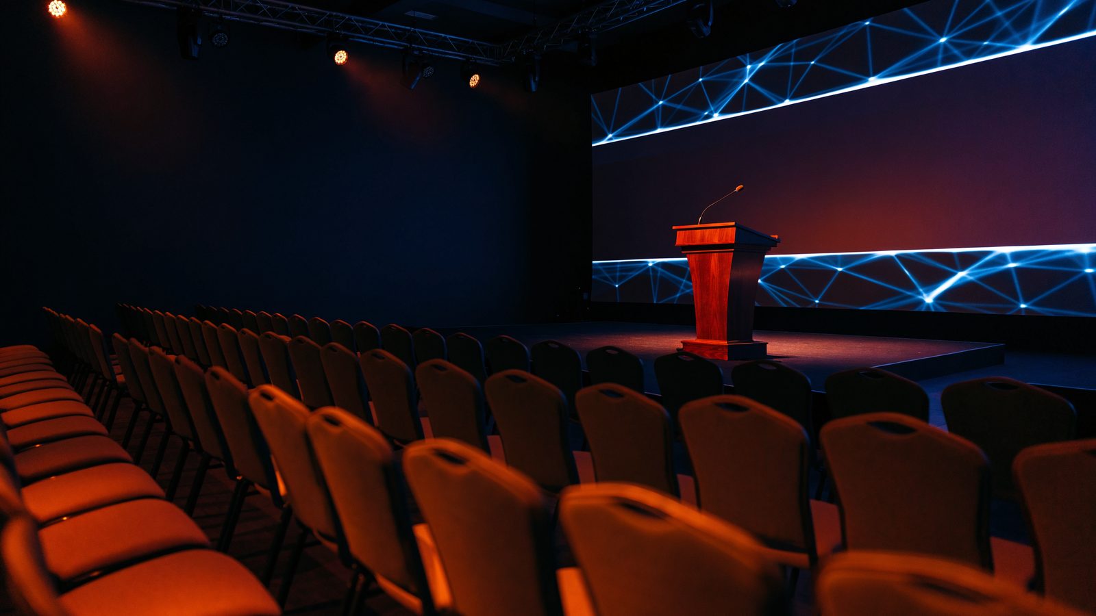 An empty conference ballroom before a keynote, with a lectern and microphone on stage, a glowing blue network pattern on the screen and rows of empty chairs.